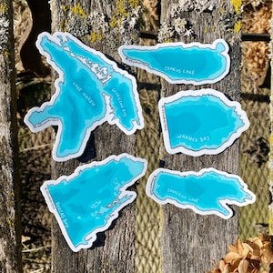 May include: Five blue and white stickers with outlines of different bodies of water. The stickers are labelled "Lake Huron", "Georgian Bay", "Cyprus Lake", "Warner Bay", and "Cameron Lake".