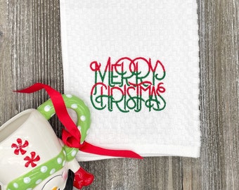 May include: White Christmas-themed kitchen towel with the embroidered words "MERRY CHRISTMAS" in red and green. A snowman mug with a red ribbon, a candle, and pine cones are also in the image.