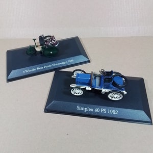 May include: Two miniature model cars on black display bases. The first car is a green 3-wheeled car with the text "3 Wheeler Benz Patent Motorwagen 1886". The second car is blue with the text "Simplex 40 PS 1902".
