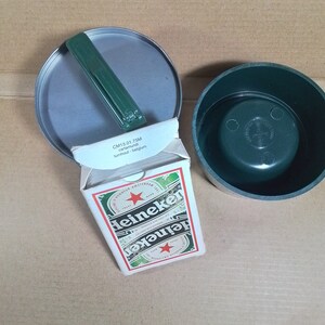 May include: A deck of playing cards in a green and white box with the Heineken logo. The box is open and the cards are visible. The box is sitting on a brown surface. The text on the box reads "CM13.01.75M cartanundi turnhout - belgium".