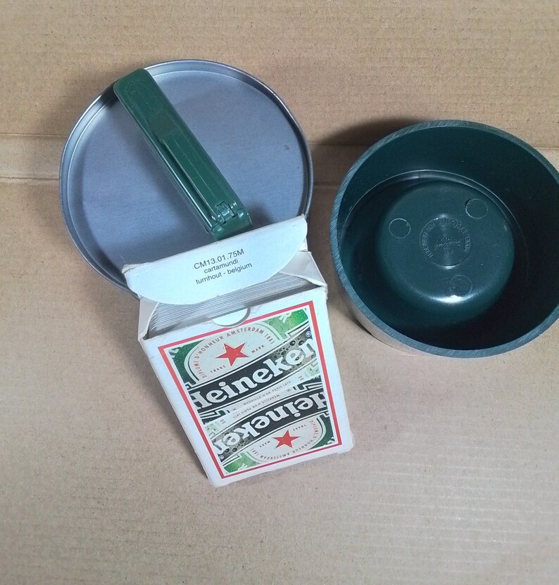 May include: A deck of playing cards in a green and white box with the Heineken logo. The box is open and the cards are visible. The box is sitting on a brown surface. The text on the box reads "CM13.01.75M cartanundi turnhout - belgium".