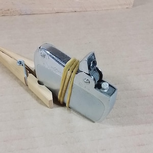 May include: A silver metal lighter secured with a yellow rubber band and a wooden clothespin. The lighter has a rectangular shape with a hinged top and a small nozzle. The clothespin is made of light-colored wood.