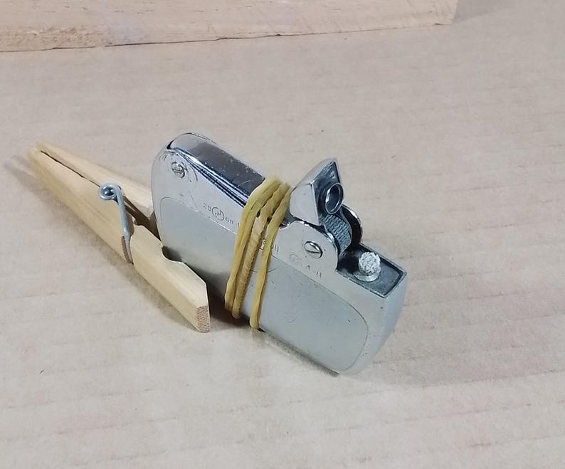 May include: A silver metal lighter secured with a yellow rubber band and a wooden clothespin. The lighter has a rectangular shape with a hinged top and a small nozzle. The clothespin is made of light-colored wood.