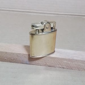 May include: A vintage, gold-toned metal lighter with a silver-colored top. The lighter has a rectangular body with rounded edges and is resting on a light-colored wooden surface. The top features a hinged mechanism for ignition.