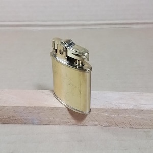 May include: A vintage gold-toned metal lighter with a rectangular body and rounded edges. The top features a textured design and a hinged lid. The lighter is resting on a light-colored wooden surface.