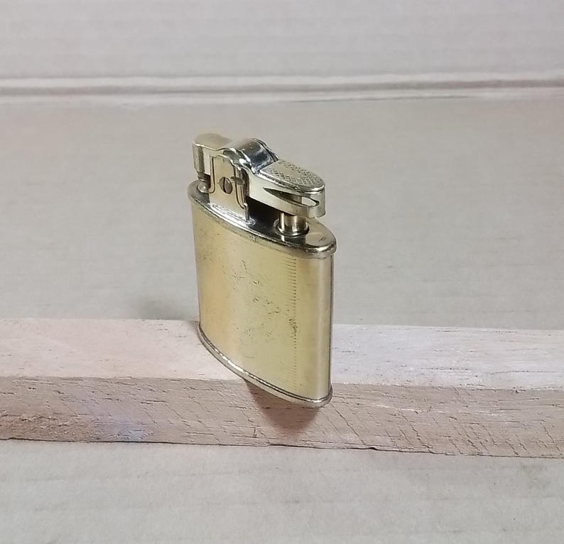 May include: A vintage gold-toned metal lighter with a rectangular body and rounded edges. The top features a textured design and a hinged lid. The lighter is resting on a light-colored wooden surface.