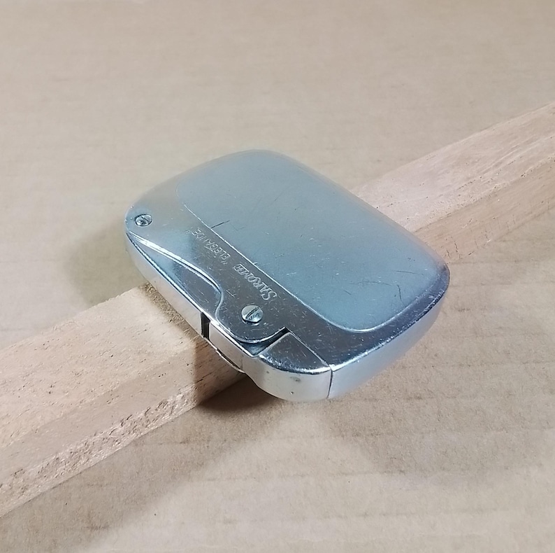May include: A silver-colored, rectangular lighter with rounded edges. The lighter has a hinged lid and a small, curved section on one side. The text "SAROME ELEGANCE" is printed on the side. The lighter is resting on a wooden surface.