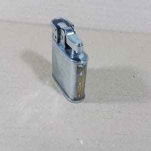 May include: A vintage silver-toned metal lighter with a rectangular body and a hinged top. The lighter shows signs of wear, with some discoloration on the edges. The top features a textured, silver-colored ignition mechanism.