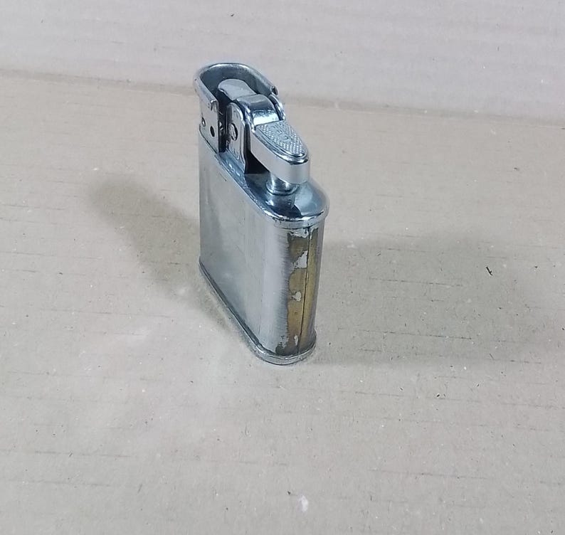 May include: A vintage silver-toned metal lighter with a rectangular body and a hinged top. The lighter shows signs of wear, with some discoloration on the edges. The top features a textured, silver-colored ignition mechanism.