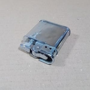 May include: A vintage silver-toned lighter with a rectangular body and rounded edges. The lighter features a textured wheel and a hinged lid. The word "FIELD" is visible on the side. The lighter is resting on a light brown surface.