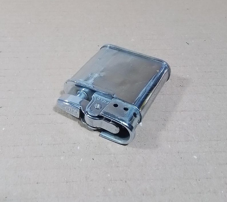 May include: A vintage silver-toned lighter with a rectangular body and rounded edges. The lighter features a textured wheel and a hinged lid. The word "FIELD" is visible on the side. The lighter is resting on a light brown surface.