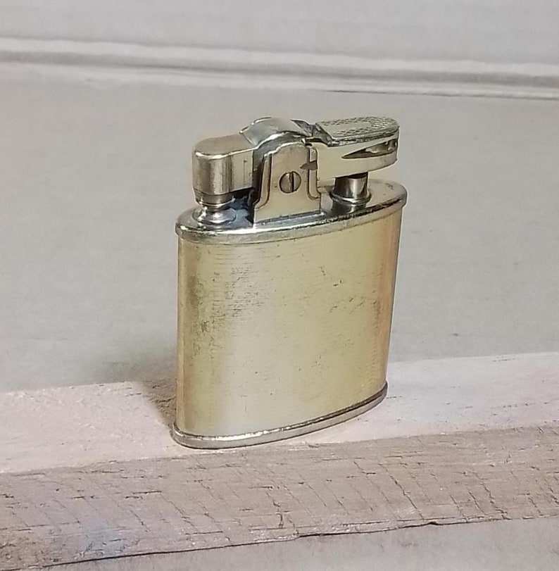 May include: A vintage, gold-toned metal lighter with a rectangular body and rounded edges. The top features a textured, silver-colored ignition mechanism. The lighter is positioned on a light-colored wooden surface.