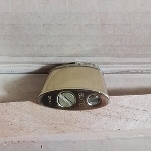 May include: A vintage silver-toned lighter with a rounded rectangular shape. The lighter has the word "BUXTON" printed on the side and "JAPAN" on the bottom. It features a screw and a small circular opening.