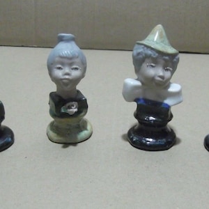 Puede incluir: Cuatro figuritas de cerámica de niños con diferentes peinados y ropa. Cada figurita está sobre una base negra.