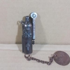 May include: An antique metal lighter with a chain and a tag. The lighter is dark gray with an arrow and decorative elements. The tag reads "1828".