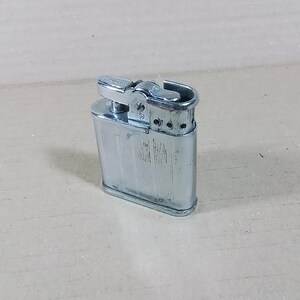 May include: A vintage silver-colored lighter with a rectangular body and a hinged top. The lighter has a textured, vertically-lined pattern on the front and a small lever for ignition. The design is classic and functional.