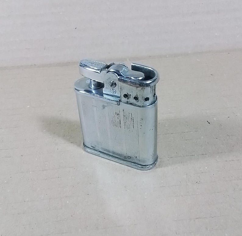 May include: A vintage silver-colored lighter with a rectangular body and a hinged top. The lighter has a textured, vertically-lined pattern on the front and a small lever for ignition. The design is classic and functional.