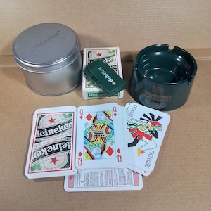 May include: A green Heineken ashtray with a silver tin can, a deck of playing cards, and a green lighter with the word "Heineken" printed on it.