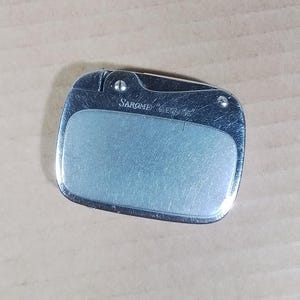 May include: A silver-toned lighter with a rectangular shape and rounded edges. The lighter has a brushed metal finish and a lighter blue center panel. The text "SAROME ELEGANCE" is printed on the top.