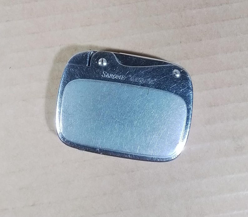 May include: A silver-toned lighter with a rectangular shape and rounded edges. The lighter has a brushed metal finish and a lighter blue center panel. The text "SAROME ELEGANCE" is printed on the top.