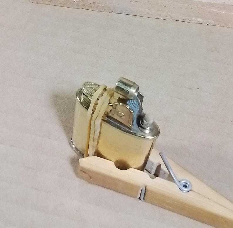 May include: A gold-colored lighter secured with a rubber band and held by a wooden clothespin. The lighter has a metallic finish and a blue accent near the ignition. The clothespin is made of light-colored wood.