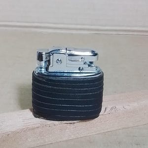 Puede incluir: Un encendedor cromado con una base negra texturizada. El encendedor tiene un diseño abatible y una base cilíndrica estriada. El encendedor se muestra sobre una superficie de madera.