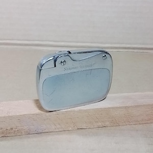 May include: A silver-toned Sarome "Elegance" lighter. The lighter has a rounded rectangular shape with a hinged top and a textured surface. The text "Sarome Elegance" is printed on the front of the lighter.