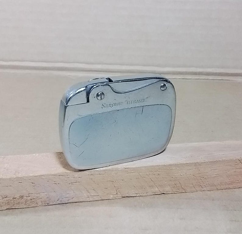 May include: A silver-toned Sarome "Elegance" lighter. The lighter has a rounded rectangular shape with a hinged top and a textured surface. The text "Sarome Elegance" is printed on the front of the lighter.