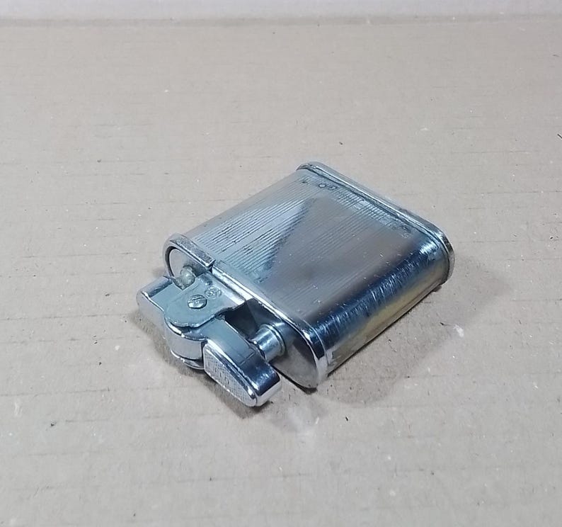 May include: A silver-colored, rectangular lighter with a textured, ridged surface. The lighter has a hinged lid and a side lever for ignition. The design is classic and functional, suitable for everyday use.