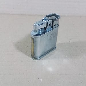 May include: A vintage silver-toned metal lighter with a textured top and a rectangular body. The lighter shows signs of wear, with some discoloration and scratches. The design is classic and functional, with a hinged top and a flint wheel.
