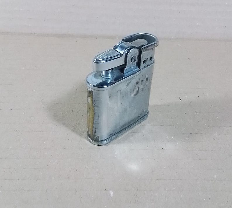 May include: A vintage silver-toned metal lighter with a textured top and a rectangular body. The lighter shows signs of wear, with some discoloration and scratches. The design is classic and functional, with a hinged top and a flint wheel.