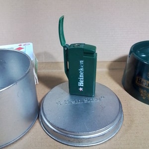 May include: Green Heineken lighter with a flip-top lid. The lighter is sitting on a silver metal coaster with the Heineken logo.