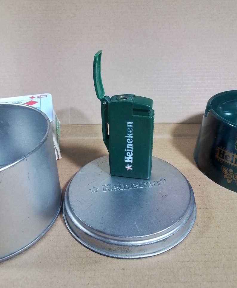 May include: Green Heineken lighter with a flip-top lid. The lighter is sitting on a silver metal coaster with the Heineken logo.