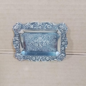 Op de afbeelding: Een rechthoekige zilverkleurige tray met sierlijke bloemen- en krulwerkdetails. Het oppervlak van de tray heeft een centraal bloemmotief en de randen zijn versierd met ingewikkelde patronen. De tray is waarschijnlijk van metaal.