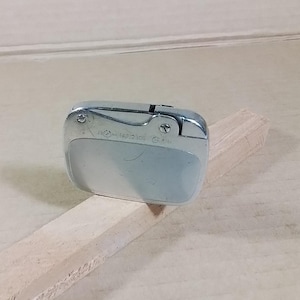 May include: A vintage silver-toned lighter with a curved design. The lighter has a rectangular shape with rounded edges and a hinged top. The surface shows some wear and tear, suggesting age. The lighter is resting on a light-colored wooden surface.