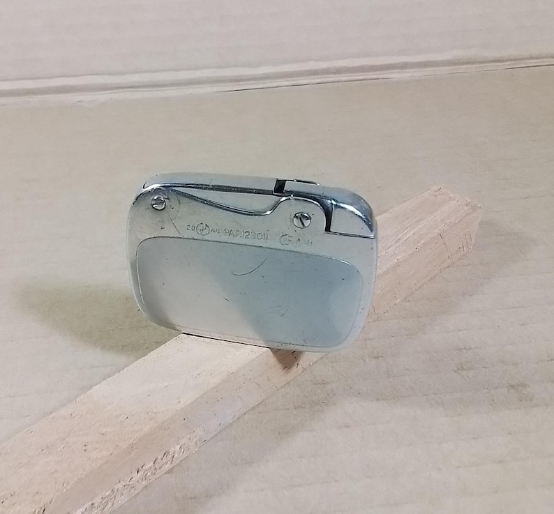 May include: A vintage silver-toned lighter with a curved design. The lighter has a rectangular shape with rounded edges and a hinged top. The surface shows some wear and tear, suggesting age. The lighter is resting on a light-colored wooden surface.