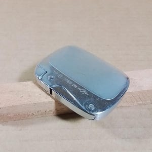 May include: A silver-toned lighter with a rectangular shape and a rounded top. The lighter has a hinged lid and a metallic finish. The text "28 ee PAT.129011, Gf--" is visible on the side. The lighter is placed on a wooden surface.