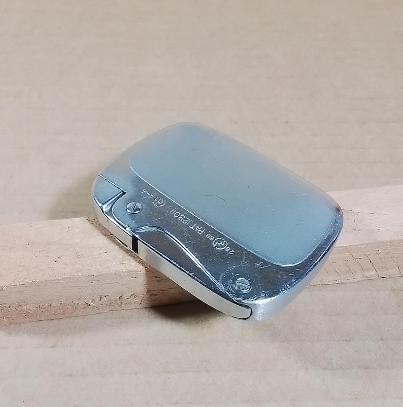 May include: A silver-toned lighter with a rectangular shape and a rounded top. The lighter has a hinged lid and a metallic finish. The text "28 ee PAT.129011, Gf--" is visible on the side. The lighter is placed on a wooden surface.