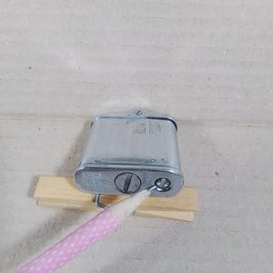 May include: A vintage silver-toned metal lighter with a rectangular body and a textured surface. The lighter has a hinged top and a circular dial on the bottom. It rests on a small wooden support.