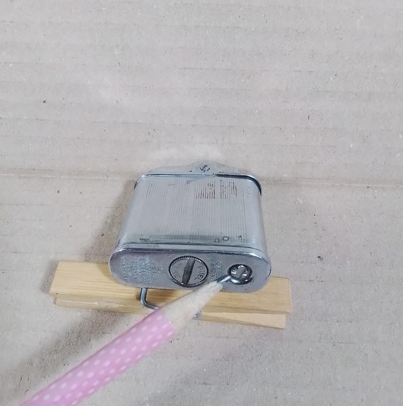 May include: A vintage silver-toned metal lighter with a rectangular body and a textured surface. The lighter has a hinged top and a circular dial on the bottom. It rests on a small wooden support.
