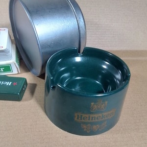 May include: Green ceramic ashtray with a gold Heineken logo. The ashtray has a deep bowl and a wide rim.