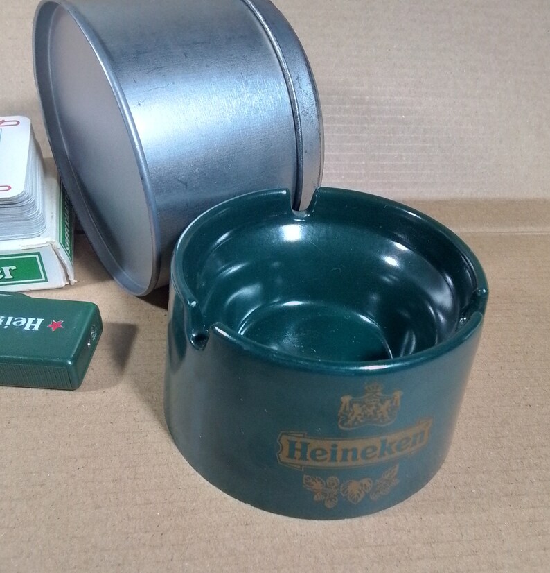 May include: Green ceramic ashtray with a gold Heineken logo. The ashtray has a deep bowl and a wide rim.
