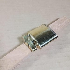 May include: A vintage brass lighter with a rectangular body and hinged lid, resting on a light-colored wooden stick. The lighter has a classic design with a flint wheel and fuel port, showcasing a polished finish.