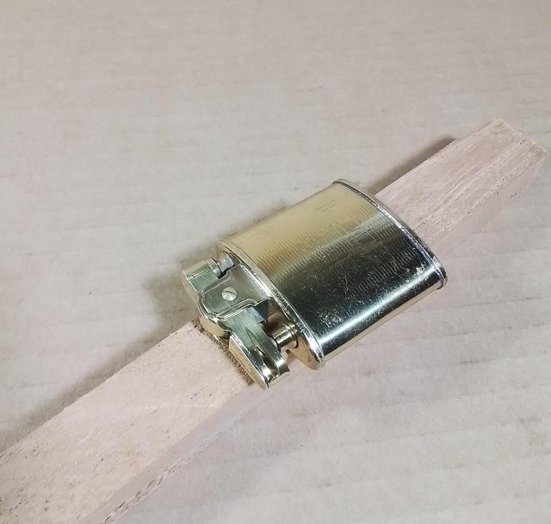 May include: A vintage brass lighter with a rectangular body and hinged lid, resting on a light-colored wooden stick. The lighter has a classic design with a flint wheel and fuel port, showcasing a polished finish.