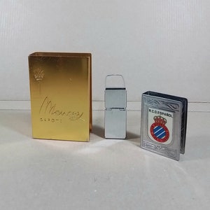Puede incluir: Tres porta-cerillas vintage. Uno es dorado con la palabra "Meneses" y el texto "Expo '71". Otro es plateado con una tapa con bisagras. El tercero es plateado con una imagen en blanco y negro de un escudo con el texto "R.C.D. ESPANOL".