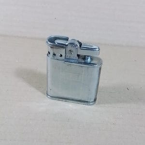 May include: A vintage silver-toned metal lighter. The lighter has a rectangular body with rounded edges and a hinged top. The lighter has a textured surface and a small lever for ignition. The word "FIELD" is visible on the side.