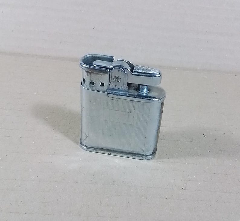 May include: A vintage silver-toned metal lighter. The lighter has a rectangular body with rounded edges and a hinged top. The lighter has a textured surface and a small lever for ignition. The word "FIELD" is visible on the side.