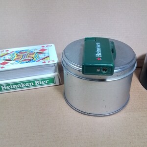 May include: A green Heineken lighter on top of a silver tin can with the text "Heineken Bier" on a deck of playing cards.
