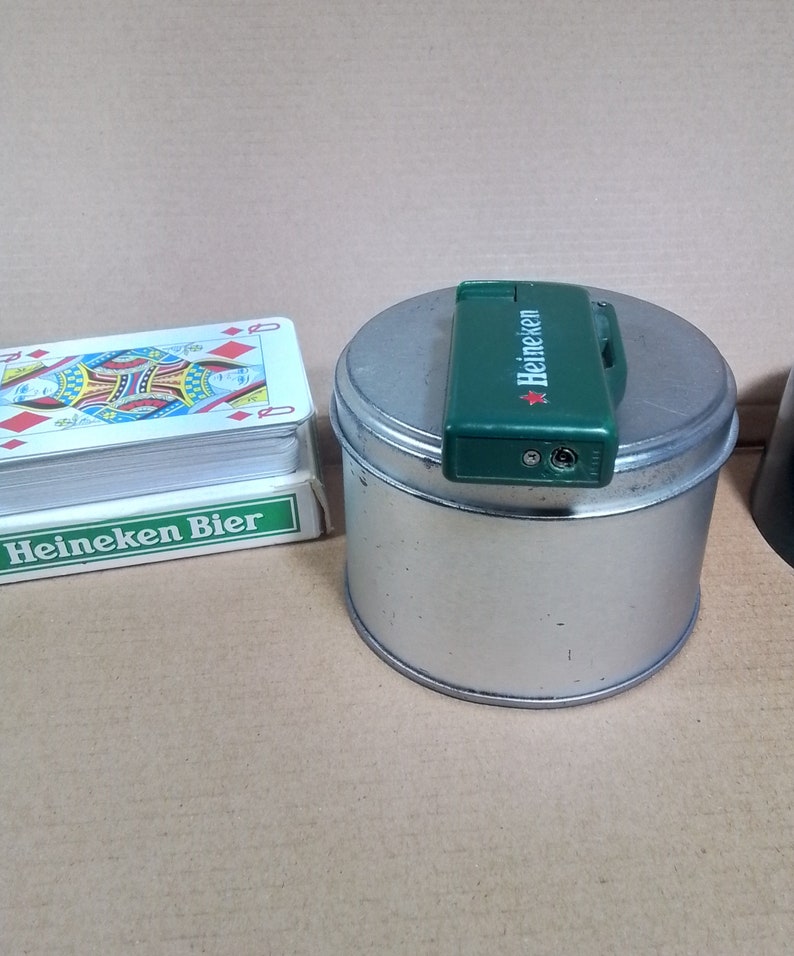 May include: A green Heineken lighter on top of a silver tin can with the text "Heineken Bier" on a deck of playing cards.