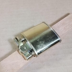 May include: A vintage, metallic gold-colored lighter with a textured surface. The lighter is rectangular with rounded edges and a hinged top. It rests on a light-colored wooden stick, showcasing its design and details.